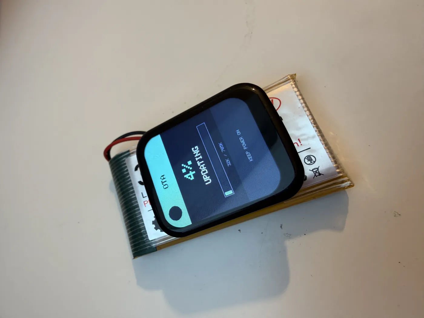 Buddy auf einem LiPo-Akku, das Display zeigt ein laufendes Firmware-Update über die OTA-Ansicht mit Fortschrittsbalken.