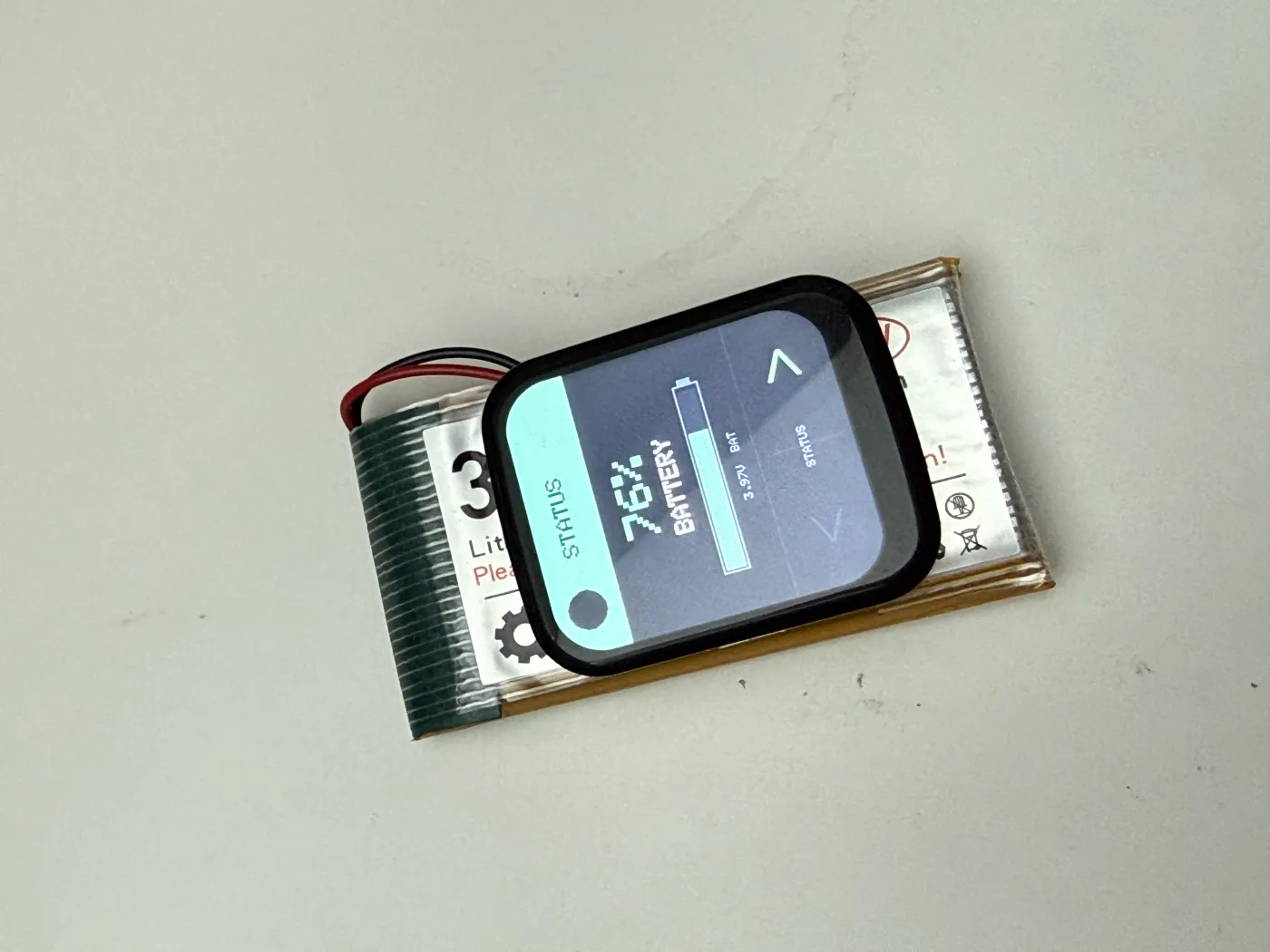 Buddy im Akkubetrieb auf einem LiPo-Akku liegend, das Display zeigt die STATUS-Ansicht mit 76 Prozent Akkustand und 3,97 Volt.
